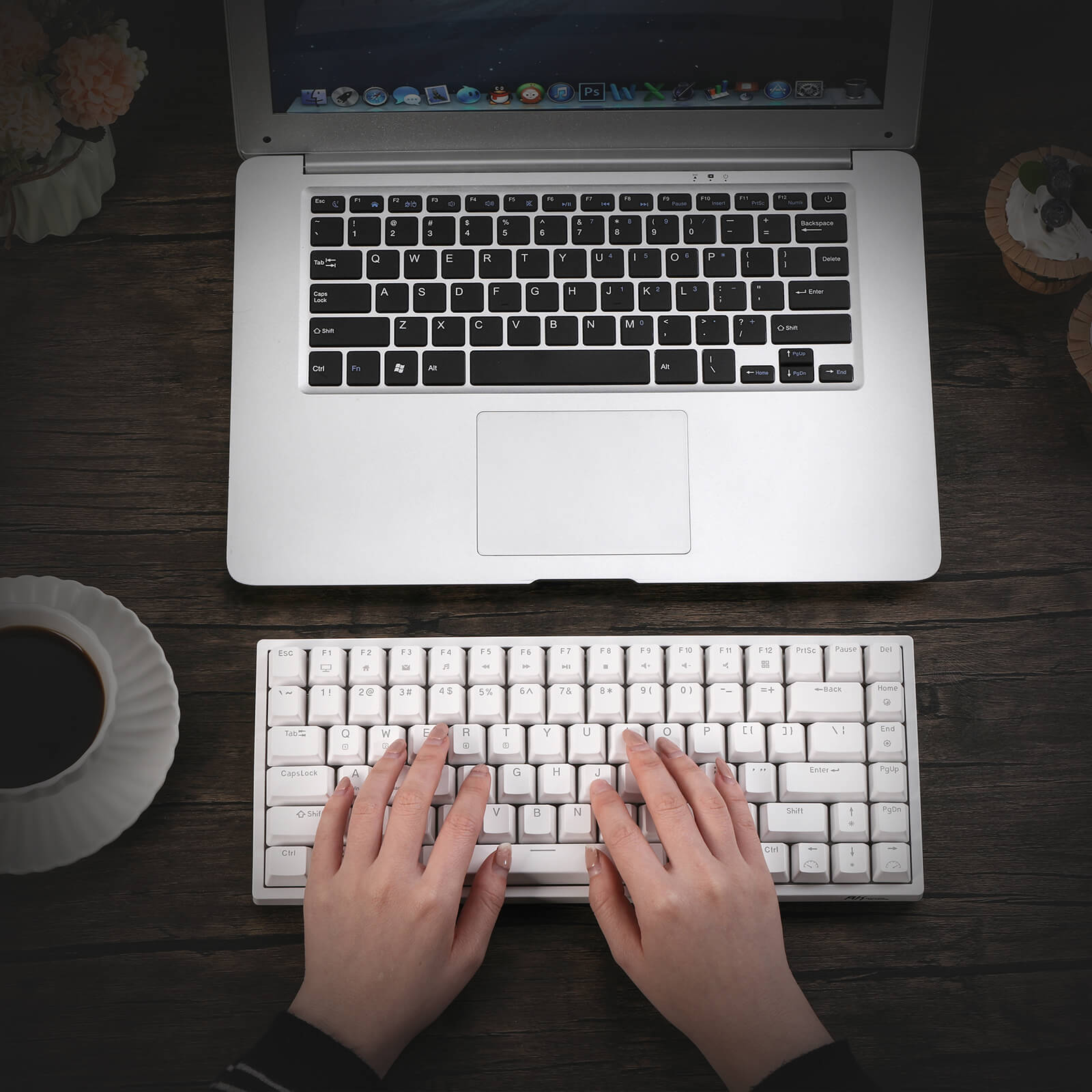 A user typing on a Royal Kludge RK84 compact gaming keyboard placed on a wooden table, alongside a laptop, a cup of coffee, and a decorative plate. The keyboard features a sleek white design and is positioned for easy access.